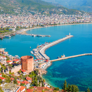 Uitzicht op de kust van Alanya met helderblauwe zee, lange havenpier, de Rode Toren en een uitgestrekte stad langs de baai.