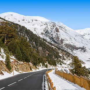 Bochtige bergweg langs besneeuwde hellingen en dennenbomen, met houten vangrail en helderblauwe lucht in de bergen.