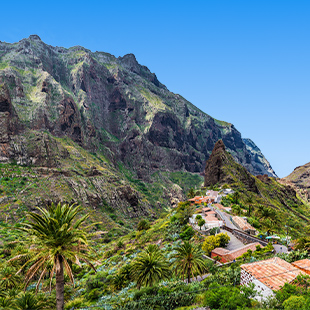 Bergdorp Masca tussen hoge rotswanden, met palmbomen, terracotta daken en kronkelende weg in een groene vallei