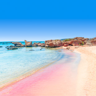 Roze strand van Elafonissi Beach met ondiep, helder zeewater, lichtroze zand en lage rotsen langs de rustige kustlijn.