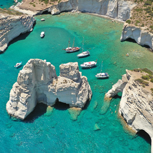 Witte rotsformaties bij Kleftiko, omringd door helder turquoise water, met meerdere kleine boten die tussen de rotsen varen.