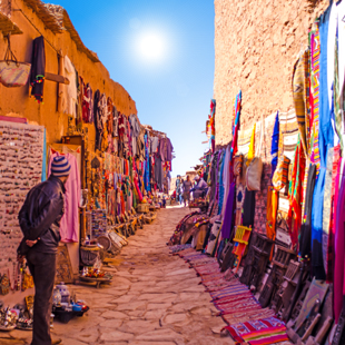 Drukke souk in Marrakech met smalle straat, kraampjes vol kleurrijke stoffen en lampen, en mensen die langs de winkels lopen.