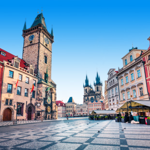 Oude stadsplein in Praag met de Astronomische Klok, historische gevels en kerkspitsen rond een leeg geplaveid plein onder een heldere blauwe lucht.