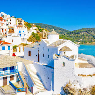 Witte huisjes op Skopelos, trapsgewijs gebouwd tegen een heuvel, met uitzicht op zee en groene bergen op de achtergrond.