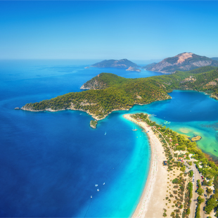 Luchtfoto van een smalle zandstrook bij een turquoise lagune en diepblauwe zee, omgeven door groene heuvels en kleine boten.