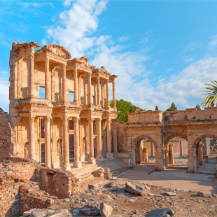 De indrukwekkende façade van de oude Bibliotheek van Celsus in Efeze, met hoge zuilen, bogen en zonverlichte ruïnes onder een blauwe hemel.