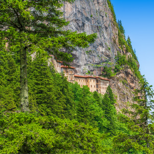 Het Sumela-klooster gebouwd tegen een steile rotswand, omringd door dicht groen bos in een bergachtig landschap.